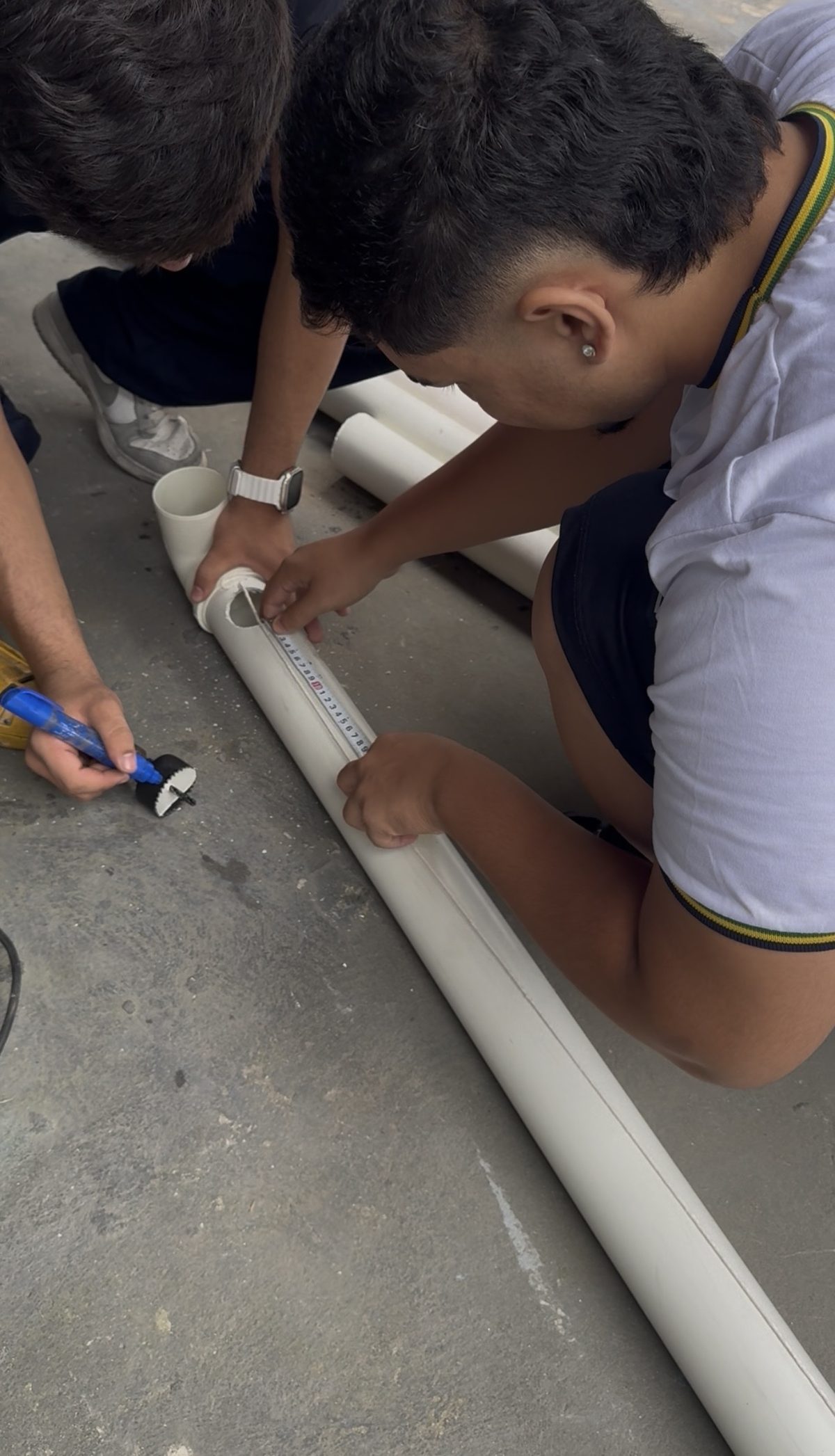 Estudantes medindo o espaçamento dos furos no tubo de PVC com trena e marcando com caneta