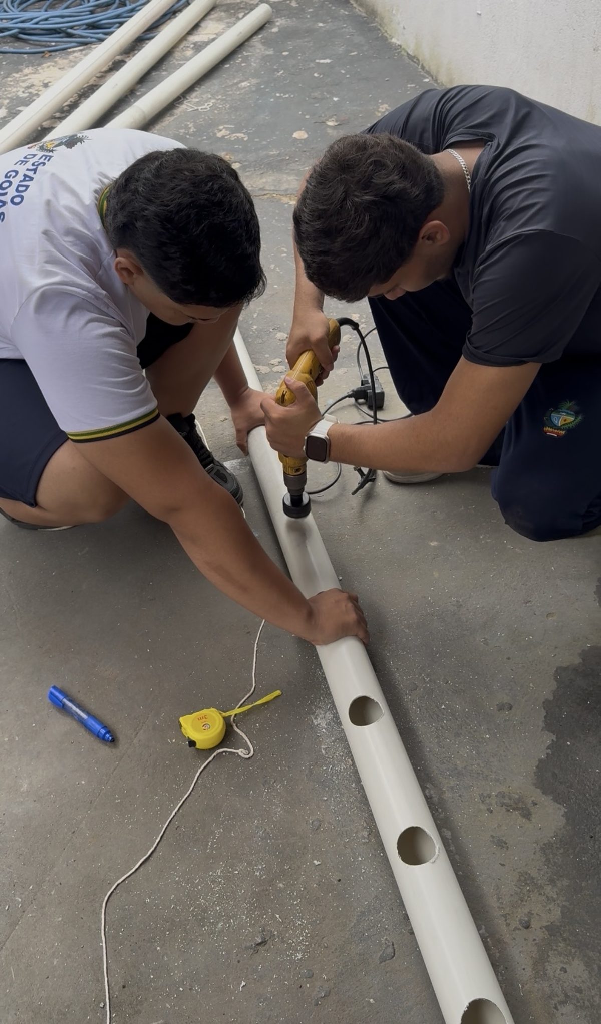 Dois estudantes perfurando o tubo de PVC com furadeira e serra copo, com trena e caneta de marcação ao lado