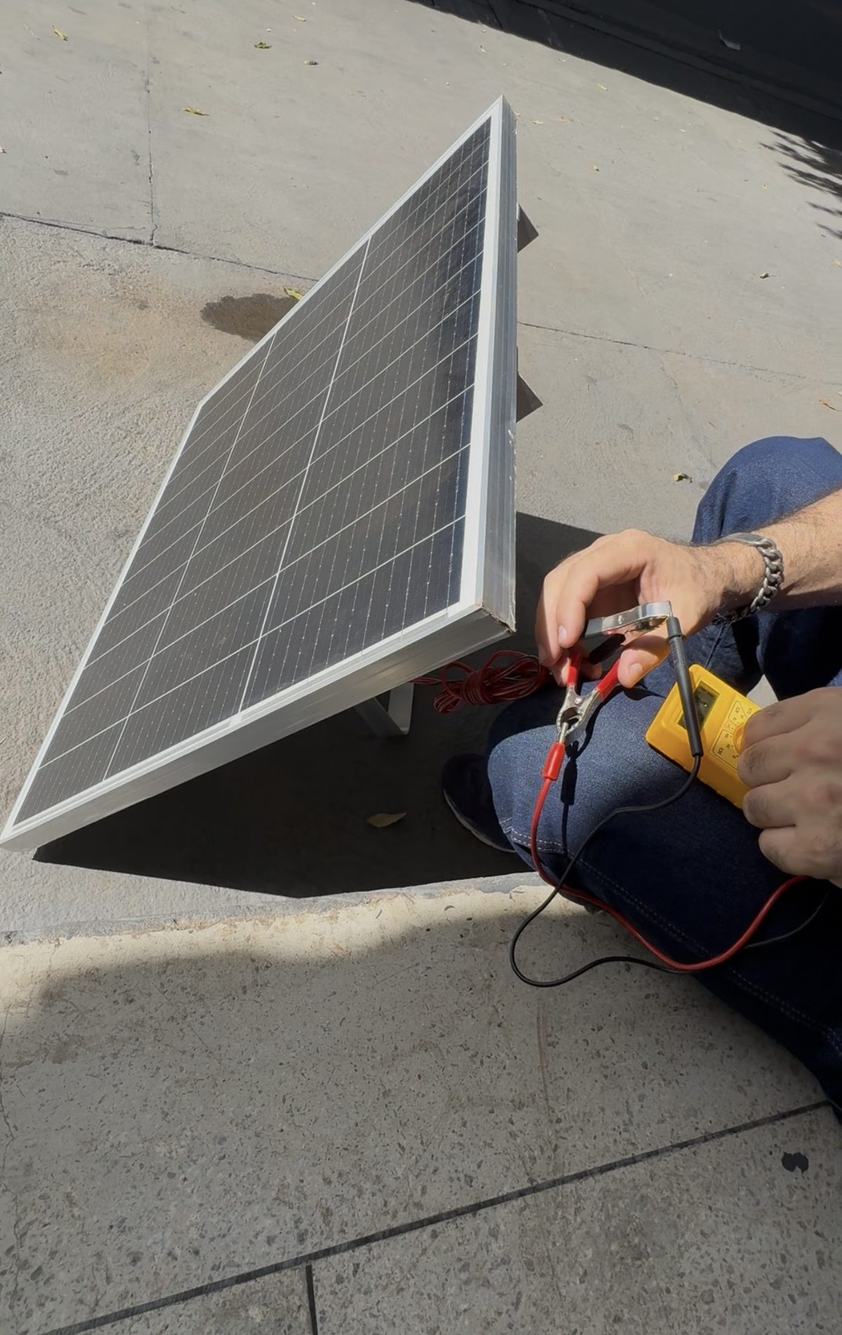 Painel solar apoiado no chão sendo testado com multímetro para medir a tensão gerada